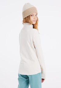 Licht beige lange mouw top met een hoge kraag, zachte textuur en een klein logo op de achterkant. Draag het met turquoise broek en een beige gebreide beanie.