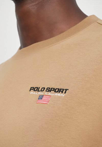 Dzeltena apģērba kreklu džemperis, kas izgatavots no mīksta auduma, ar melnām izšūtām uzrakstiem "POLO SPORT" un "RALPH LAUREN", kā arī nelielu ASV karoga akcentu.