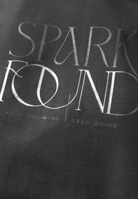 Tumma harmaa kangas, jossa metallinhohtoinen hopeateksti "SPARK FOUND" ja "KEEP GROWING | KEEP GOING", jonka pinnassa on sileä pinta ja vahva typografia.
