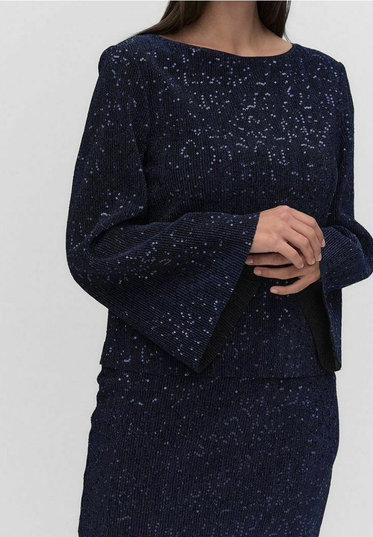 Femme portant un ensemble en maille côtelée bleu marine composé d'un haut et d'une jupe, avec des manches longues évasées et des paillettes brillantes parsemées.