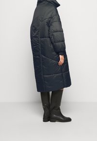 Cappotto imbottito blu navy con colletto alto, caratterizzato da cuciture diagonali e polsini elasticizzati, indossato con stivali neri alti.
