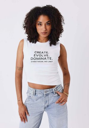 Vrouw met krullend haar, die een wit mouwloos topje met tekst "Create. Evolve. Dominate." en lichtblauwe spijkerbroek draagt tegen een effen achtergrond.