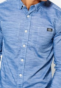Jasnoniebieska koszula z długim rękawem i kołnierzykiem zapinanym na guziki, posiadająca dwie kieszenie na piersi oraz małą czarną naszywkę z logo na lewej kieszeni.