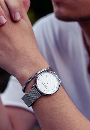 Montre avec bracelet en maille argentée, affichage blanc, design minimaliste et marqueurs argentés. Accompagnée d'un bracelet métallique fin.