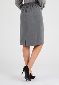 Jupe grise droite avec une taille élastique, longueur genou, présentant une fente au dos. Portée avec des chaussures à talons hauts noires. Texture unie.