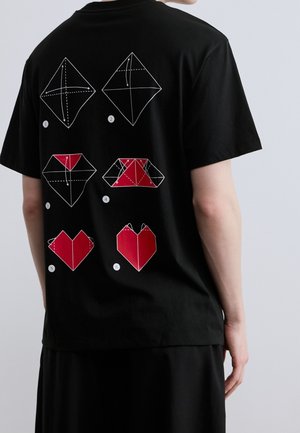 Osoba ubrana w czarną koszulkę z sześcioma białymi i czerwonymi ilustracjami kroków składania origami w kształcie serca na plecach na jasnoszarym tle.