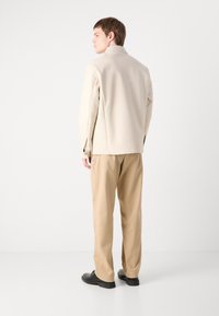 Beige tailored jacket made of smooth fabric, paired with light beige trousers. Black shoes complete the outfit. Clean, minimalist design.