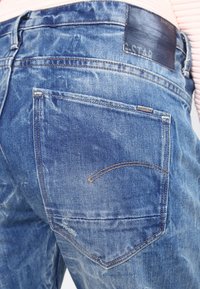 Jeans de mezclilla azul desgastados con un diseño clásico de cinco bolsillos. Presentan un parche de cuero en la cintura y sutiles acentos de costura.