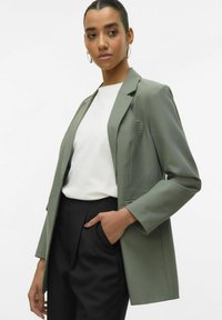 Blazer vert sur mesure en tissu lisse et léger, avec un détail de poche unique, porté sur un haut blanc et un pantalon noir.