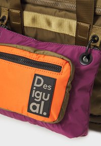 Bolsa multicolorida con un bolsillo morado y naranja, con un cierre negro y etiqueta de logo, fabricada en un tejido ligero y texturizado.