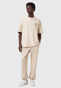Ung mand står i en beige oversized t-shirt i 100% bomuld og matchende joggers med hvide sneakers på grå baggrund.