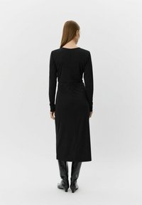 Kvinde med langt hår iført en tætsiddende, sort midikjole med lange ærmer og sorte hæleboots, stående foran en ensfarvet hvid baggrund.