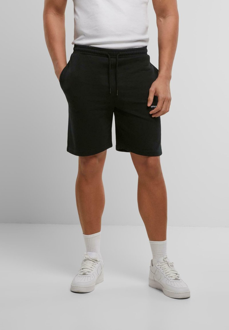 Shorts en coton noirs avec une taille élastique et un cordon de serrage ; poches sur les côtés ; associés à des baskets blanches et des chaussettes blanches de longueur mi-mollet.