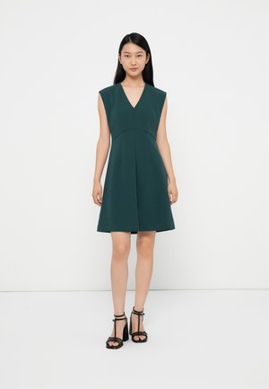 Green, sleeveless dress with a V-neck, flared skirt, and structured bodice; worn with black strappy heels; smooth texture.