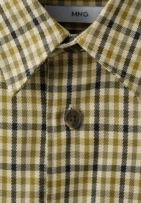 Chemise à carreaux beige et vert olive avec des lignes noires, boutonnée avec un bouton brun foncé à quatre trous, accrochée sur un cintre étiqueté "MNG".