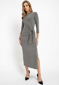 Graues, gestricktes Midi-Kleid mit Wickeldesign, drei Viertelärmel, einem seitlichen Knoten und einem Oberschenkel-spalt, kombiniert mit schwarzen High Heels.