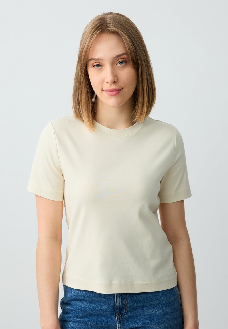 Beige T-shirt met korte mouwen, een ronde hals en een getailleerd ontwerp, gemaakt van een zachte, lichte stof; gecombineerd met blauwe jeans.
