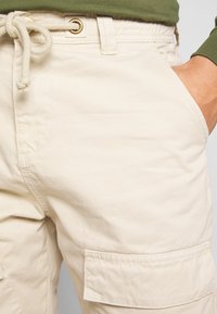 Beige cargo-housut, joissa on kiristysvyötärö, sivutaskut ja läpätasku reidessä. Valmistettu pehmeästä ja kestävästä kankaasta, jossa on sileä rakenne.