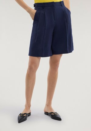 Osoba ubrana w granatowe plisowane spodenki sięgające do kolan, żółtą bluzkę, z rękami w kieszeniach oraz czarne wsuwane buty z ostrym noskiem.