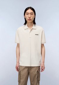 Cremefarbenes Poloshirt mit kurzen Ärmeln, Kragen und einem kleinen Logo-Patch auf der linken Brust. Kombiniert mit khakifarbenen Cargohosen. Glatte Stoffstruktur.