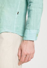 Camicia di lino turchese chiaro con una texture liscia, maniche lunghe e polsini abbottonati. Dettagli significativi della targhetta laterale e dell'orlo curvo.