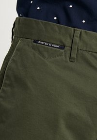 Olivgrüne Baumwollhose mit einer Vordertasche und einem Etikett, auf dem "Scotch & Soda" in Weiß auf einem marineblauen Hintergrund steht. Geschmeidige Textur.