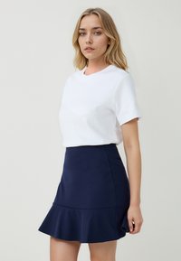 Jonge vrouw met blond haar draagt een witte top met korte mouwen en een marineblauwe uitlopende minirok tegen een lichte achtergrond.