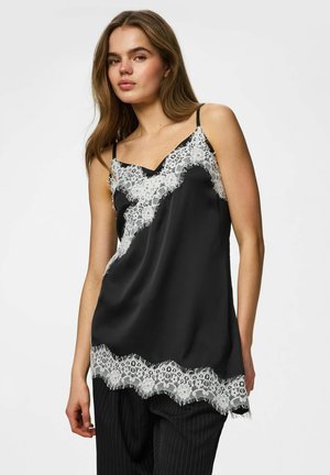 Jeune femme portant un caraco en satin noir avec bordure en dentelle blanche et pantalon noir à rayures, debout devant un fond clair uni.