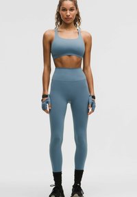 Femme debout, portant un brassard de sport bleu, un legging taille haute, des gants sans doigts et des baskets noires sur un fond uni.