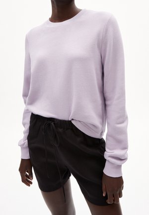 Personne portant un pull lavande à manches longues et un short noir à cordon, debout devant un fond blanc uni.