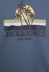 Modrá látka s grafickým designem zobrazujícím palmy proti žlutému slunci, se vrstvami zelených a černých pruhů níže. Text "JACK&JONES OAHU, HAWAI".
