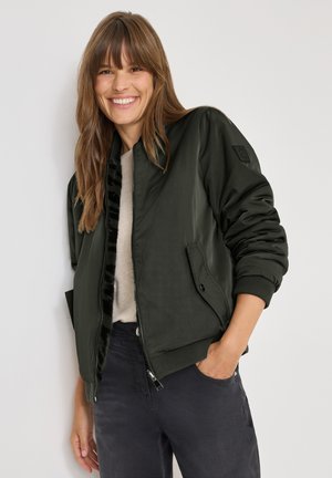 Chaqueta bomber verde oliva con cierre frontal, puños acanalados y bolsillos. Forrada con material negro suave, lleva puesta sobre un suéter claro y pantalones oscuros.
