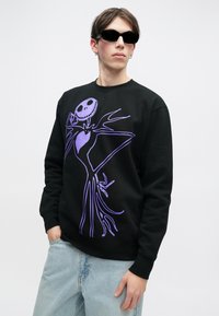 Jovem vestido com um moletom preto com uma grande estampa de esqueleto roxo, jeans azul claros, óculos de sol pretos e brincos de argola prateados, em fundo branco.