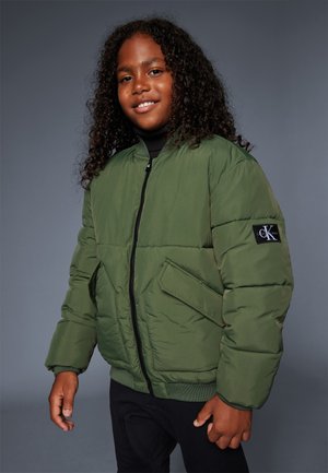 Giacca a vento verde con zip frontale e tasche laterali, caratterizzata da una patch nera con logo sul maniche. Abbinata a pantaloni neri.