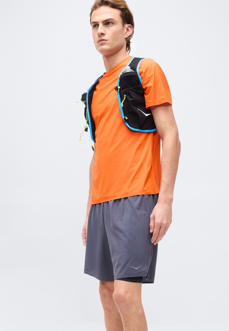 Mann trägt ein oranges Kurzarmhemd und graue Sportshorts mit einer schwarzen und blauen Trinkweste, hält Flaschen in der Hand.