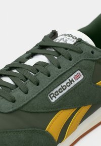Sapatilhas verdes em camurça com detalhes amarelos, atacadores brancos e patch com logotipo. Apresenta um cabedal texturizado e uma sola de borracha robusta.