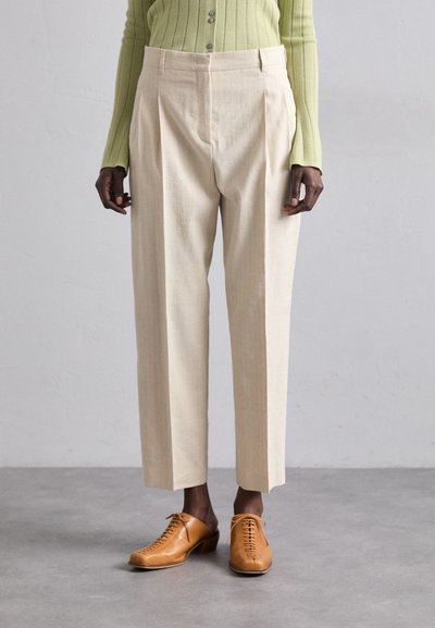 Beige, gefaltete Hose mit geradem Bein, aus strukturiertem Stoff. Kombiniert mit braunen Schnürschuhen.