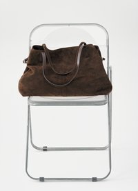 Bolso de ante marrón oscuro con correas de cuero apoyado sobre una silla plegable transparente contra un fondo blanco.