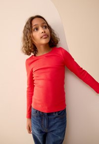 Red long-sleeve ribbed shirt, fitted design, paired with blue denim jeans. The child leans against a light-colored wall.