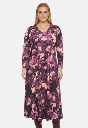 Frau trägt ein langärmeliges, violettes Blumenkleid mit V-Ausschnitt und steht vor einem schlichten weißen Hintergrund.