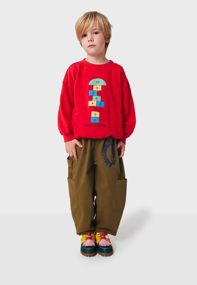 Felpa rossa con un motivo di campana, dettagli verde e giallo. Pantaloni cargo verde oliva con tasche laterali. Scarpe multicolori con fiocchi.