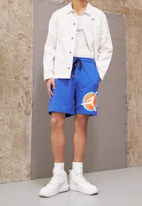 Pantaloni sportivi blu con una grafica bianca e dettagli arancioni, abbinati a una giacca bianca con bottoni e scarpe alte bianche.