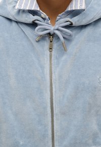 Sudadera con cremallera de color azul claro, fabricada en material suave, con capucha ajustable mediante cordones, herrajes plateados y una cremallera visible. Presenta un cuello a rayas.