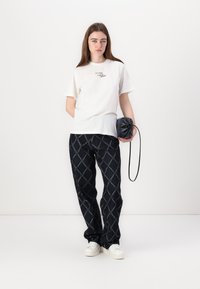 Hvit bomull T-skjorte med tekstprint, kombinert med svarte mønstrede bukser. Modellen holder en liten svart veske og har på seg hvite sneakers.