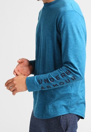 Mann trägt ein langärmeliges blaues Under Armour Shirt mit Logo und Schriftzug auf dem rechten Ärmel und steht vor einem schlichten Hintergrund.