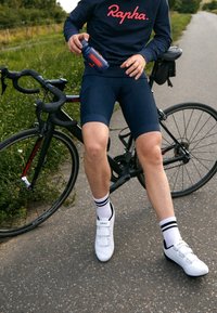 Ciclista in abbigliamento Rapha che tiene in mano una borraccia, in piedi accanto a una bici da strada nera su un sentiero asfaltato con vegetazione nelle vicinanze.