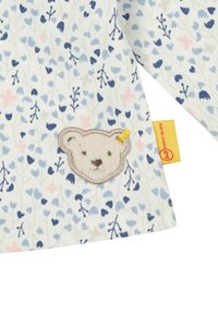 Camicia di cotone con motivo floreale in tonalità blu e rosa, con un morbido panno ricamato raffigurante la faccia di un orsetto e un'etichetta gialla in contrasto.