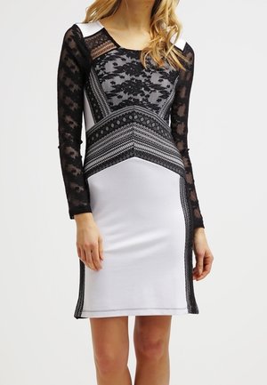 Femme portant une robe blanche ajustée avec des manches longues en dentelle noire et des motifs géométriques en dentelle sur le corsage et les côtés.