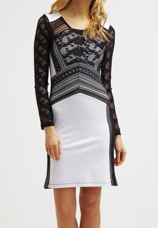 Femme portant une robe blanche ajustée avec des manches longues en dentelle noire et des motifs géométriques en dentelle sur le corsage et les côtés.