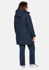 Marineblaue wasserdichte Parka mit Kapuze, gerade geschnitten, mit glatter Oberfläche und versteckten Seitentaschen. Getragen mit blauen Jeans und Sneakers.